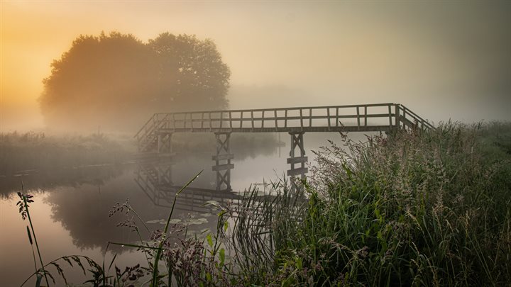 Vroeg in de ochtend komt de zon op over een mistig Hunzedal. In het midden van het beeld loopt een brug die aan de overkant van de beek in de mist verdwijnt. Op de voorgrond staat riet. De foto ademt een dromerige sfeer.